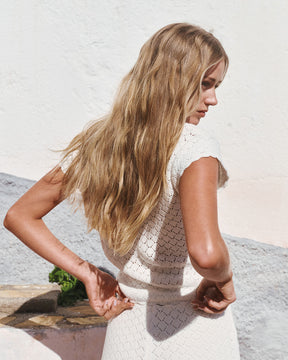 Woman with long blonde hair wearing a white sleeveless dress against a light background